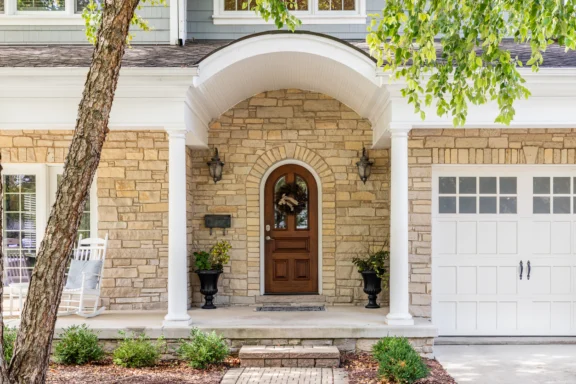 Stone front entrance with arched white porch, wooden door, and two lanterns flanking the doorway (house with a garage on the right).
