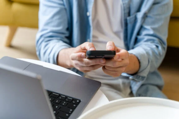 Person in a light denim jacket holds a smartphone, with a laptop nearby on a desk.