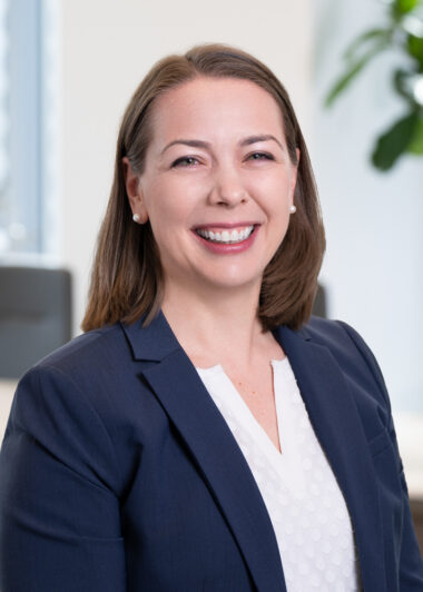 Professional portrait of a smiling woman in a navy blazer in an office setting, pearl earrings visible